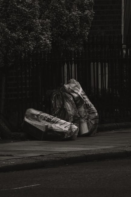 Three large black plastic rubbish bags filled with waste are placed on the edge of a street pavement in front of a black metal fence and dense foliage. The bags appear to be tightly packed and slightl